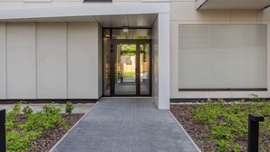 A modern building entrance with a glass door, a pathway, and a small garden area. - Bright Apartment with Parking and Balcony | Close to the Airport (Warszawa)