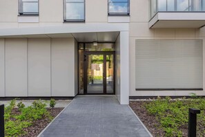 A modern building entrance with a glass door, a pathway, and a small garden area. - Bright Apartment with Parking & Balcony Close to the Airport (Warszawa)