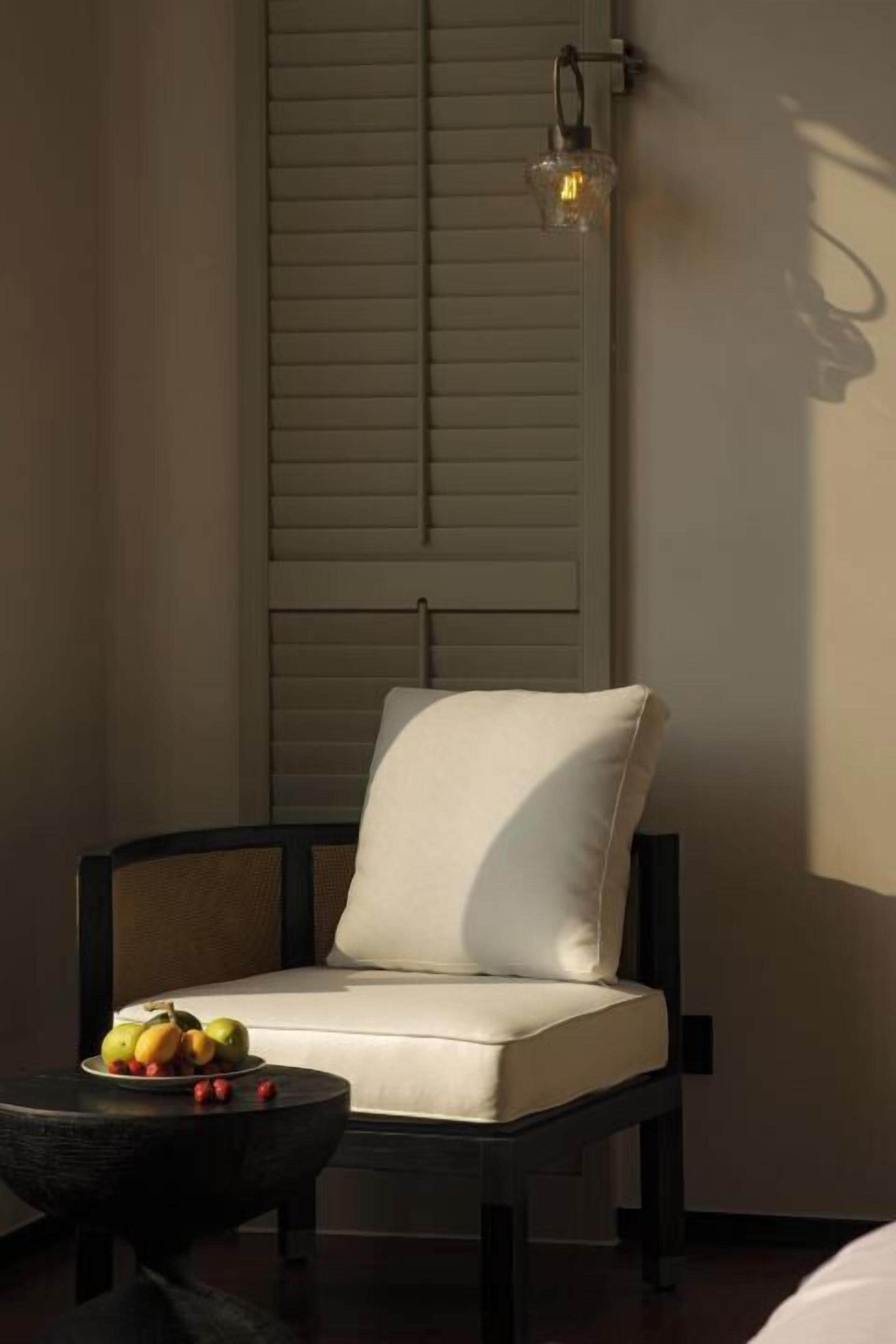 A room with a cushioned chair, a small table with fruit, and a wall-mounted light fixture.