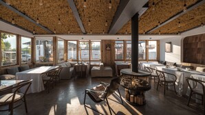 Un restaurante con techo alto, vigas de madera y grandes ventanales. Mesas y sillas dispuestas para comer.