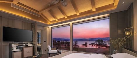 Una habitación de hotel moderna con una cama grande, un televisor de pantalla plana, un escritorio con silla y vista a un atardecer sobre el agua.