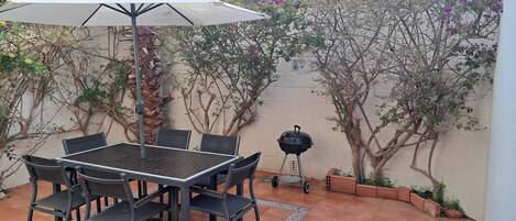 Un patio al aire libre con una mesa, sillas, una parrilla y una pérgola con un paraguas blanco.