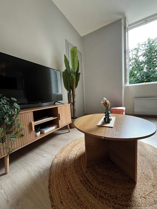 Ein modernes Wohnzimmer mit Flachbildfernseher, Holzunterhaltungseinheit, rundem Couchtisch, Topfpflanze und Blick durch ein Fenster.