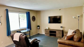 Een woonkamer met een bruine leren bank, een bruine fauteuil, een aan de muur bevestigde televisie, een bureau met lades en een raam met blauwe gordijnen.