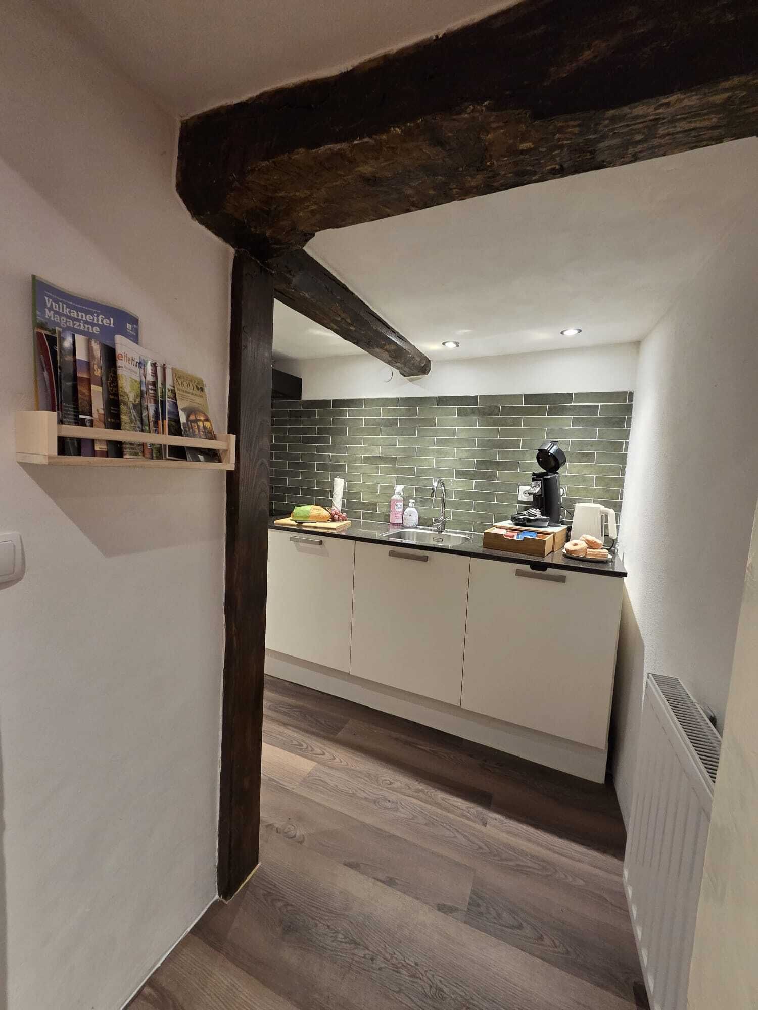 A modern kitchen with a dark wooden beam, white cabinets, a tiled backsplash, and a small shelf with magazines.