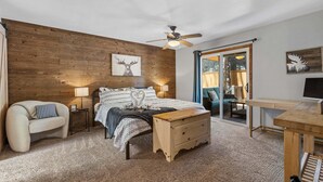 Ein Schlafzimmer mit Holzwand, einem Bett mit gemusterten Betttextilien, einem Nachttisch, einer Holzbank, einem Schreibtisch und einem Deckenventilator.