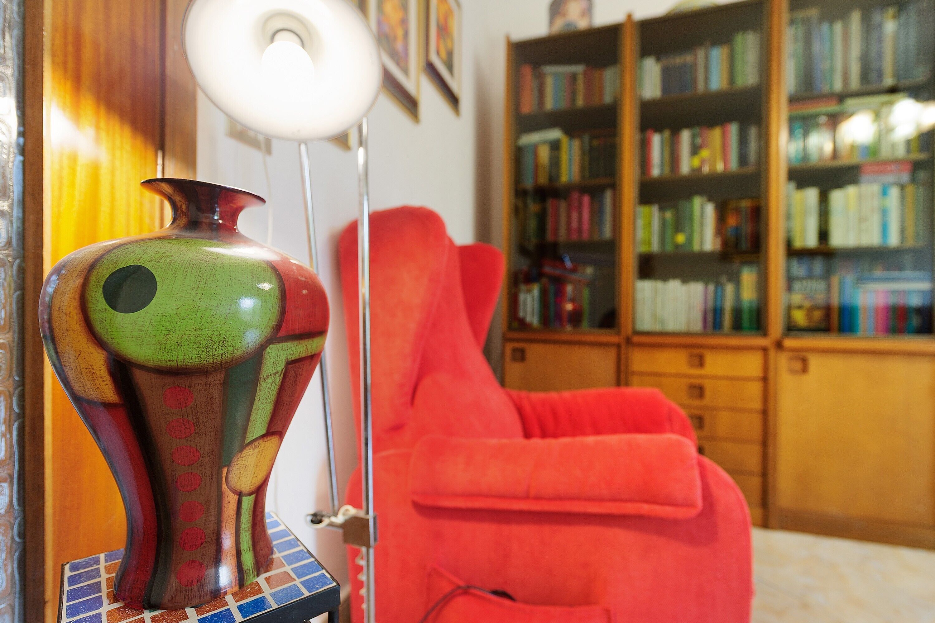 Une pièce avec une bibliothèque, un fauteuil rouge et un vase aux couleurs vives.