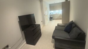 A living area with a TV, a sofa, and a kitchen in the background. - 1 Bedroom - Rubix (London)