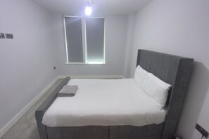 Ein kleines, ordentlich bezogenes Bett mit grauem Kopfteil und weißer Bettwäsche in einem Zimmer mit hellen Wänden und einem Fenster.