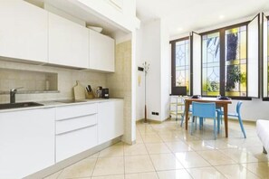 A modern kitchen with white cabinets, a sink, and a dining area with blue chairs and a table. - Hostdomus - Zoe Apartment (Alassio)