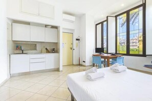 A hotel room with a bed, a desk, chairs, a kitchenette, and a view through a window. - Hostdomus - Zoe Apartment (Alassio)