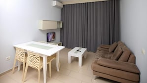 A living room with a dining table, chairs, a sofa, a TV, and a window with curtains. - Britannia Resort & SPA - All Inclusive (Vlorë)