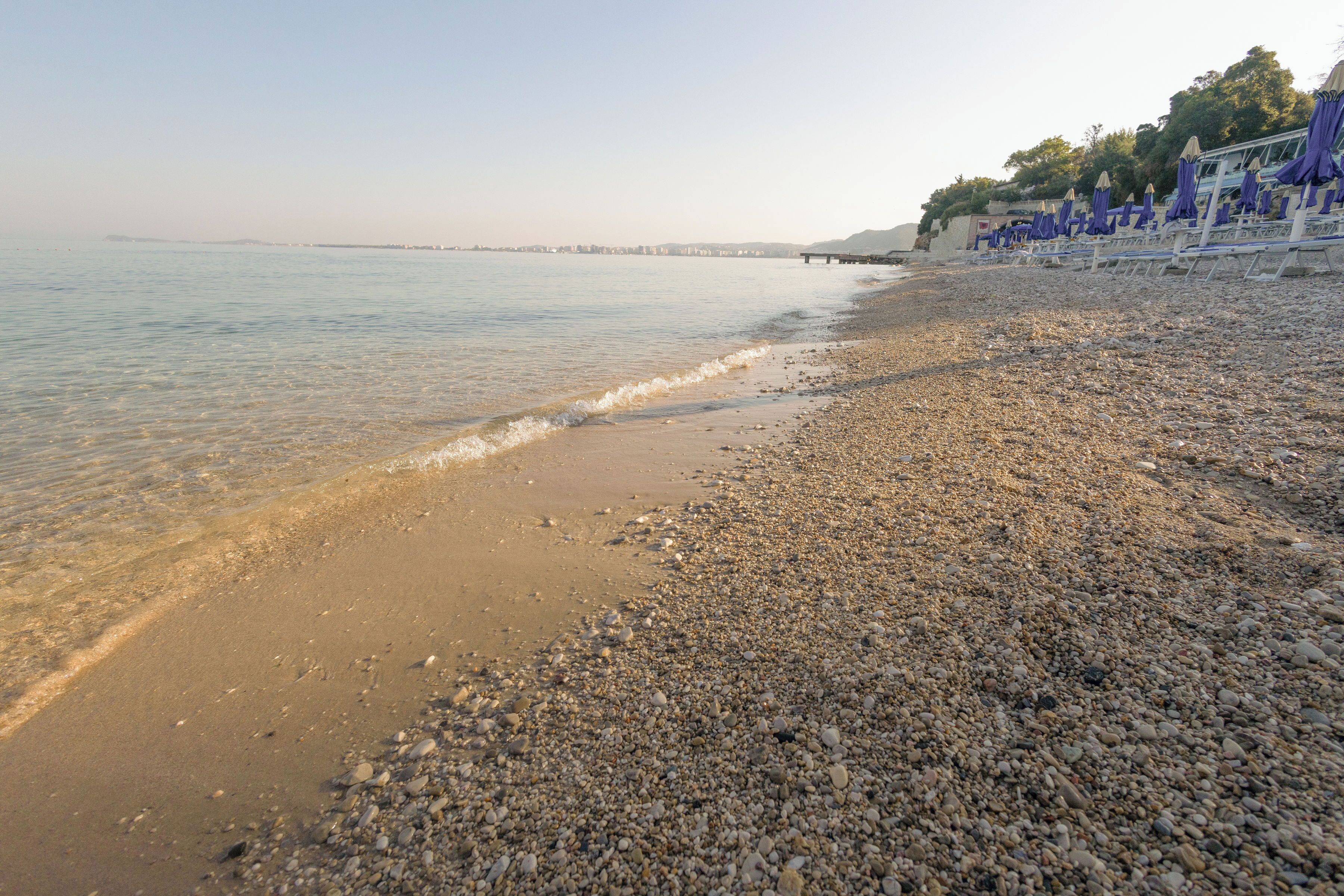 A pebbled beach with a calm sea, a distant pier, and beach umbrellas.