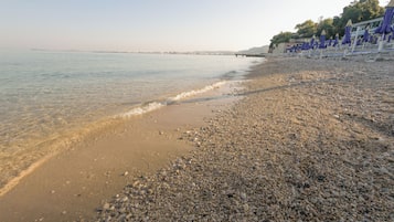 A pebbled beach with a calm sea, a distant pier, and beach umbrellas.