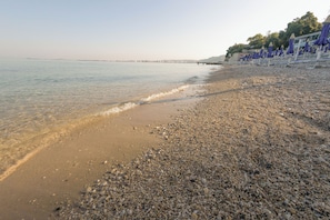A pebbled beach with a calm sea, a distant pier, and beach umbrellas. - Britannia Resort & SPA - All Inclusive (Vlorë)