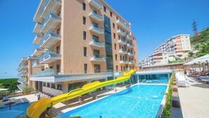 A multi-story building with balconies, a swimming pool with a yellow slide, and a clear blue sky. - Britannia Resort & SPA - All Inclusive (Vlorë)