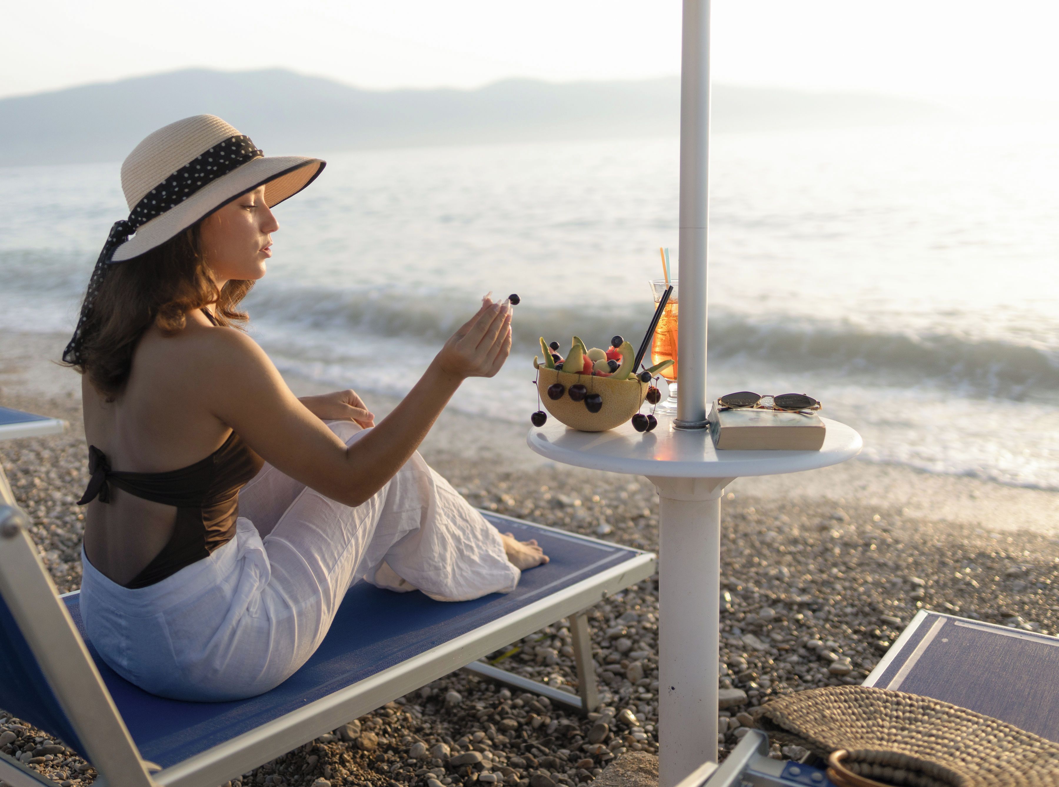 Eine Person sitzt auf einem Liegestuhl am Strand, neben sich ein Tisch mit Obstschale, Getränk und Buch.