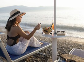 Uma pessoa sentada em uma cadeira de praia, com uma mesa ao lado contendo uma tigela de frutas, uma bebida e um livro.