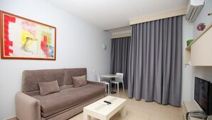 A living room with a sofa, a coffee table, a TV, a painting, a dining table, and a window with curtains. - Britannia Resort & SPA - All Inclusive (Vlorë)