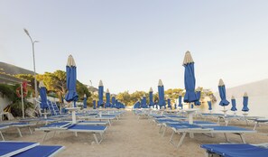 A beach with rows of sun loungers and blue umbrellas, a lifebuoy, and a clear sky. - Britannia Resort & SPA (Vlorë)