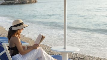 Eine Person sitzt in einem Strandstuhl unter einem blauen Sonnenschirm und liest ein Buch.