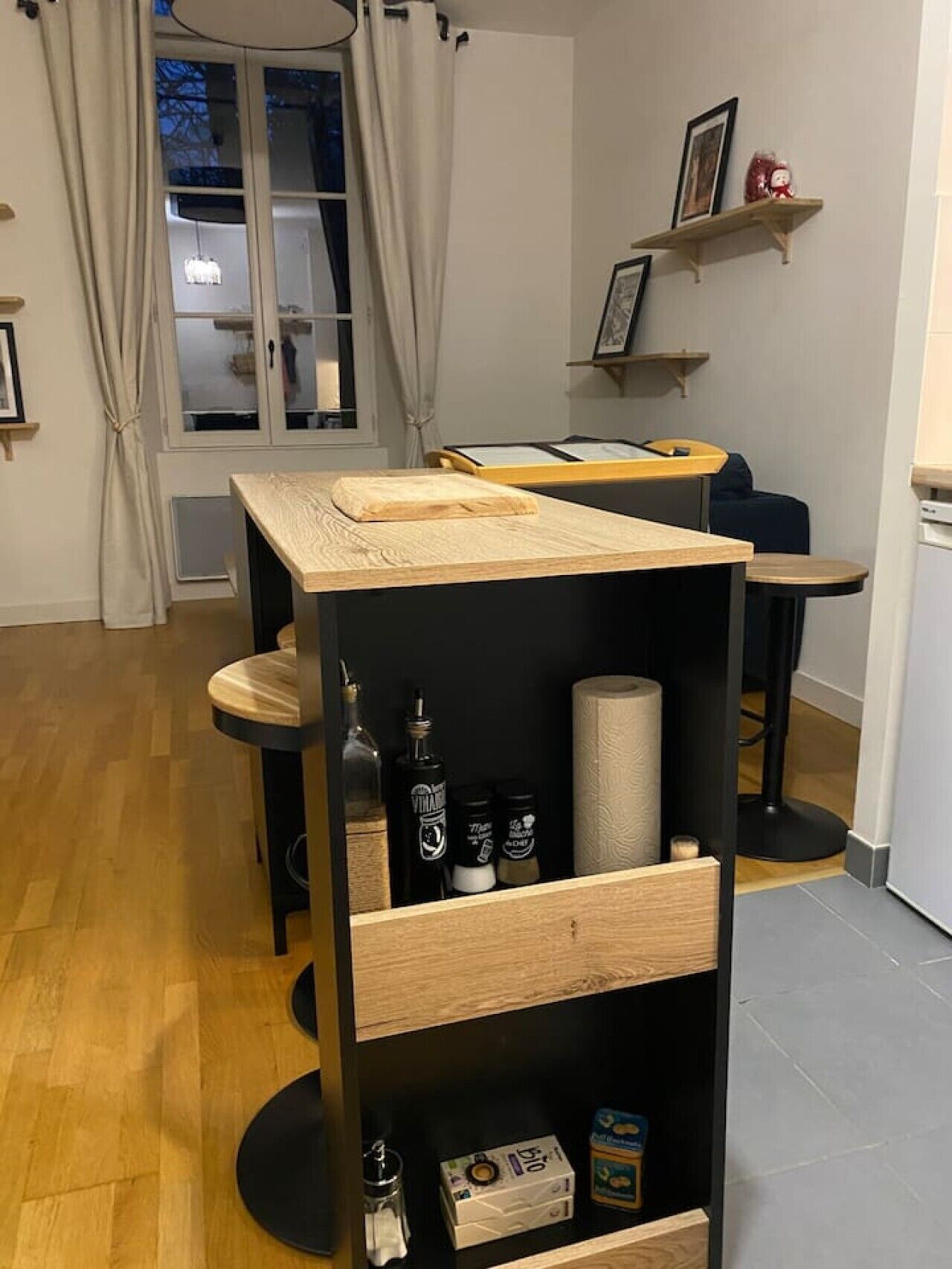 A compact room with a wooden table, a black chair, a small shelf with items, and a window with curtains.