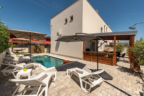 Zona de piscina con sillones de descanso, sombrillas y una pérgola de madera.