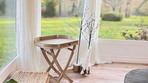 Una habitación con una mesa de madera, una silla, una planta en maceta y vistas a un jardín a través de ventanas amplias con cortinas blancas.