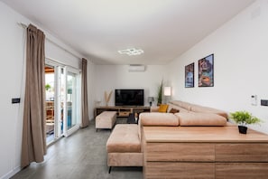 Ein modernes Wohnzimmer mit Sofa, Hocker, Fernseher und einer Schiebeglastür zum Außenbereich.
