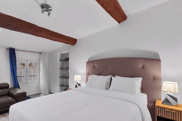 A bedroom with a large bed, a wooden ceiling beam, a nightstand, a lamp, a bookshelf, and a brown leather chair. - ☀️ In the heart of Rue Meynadier & Palais (Cannes)