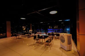 A modern indoor seating area with tables and chairs, a large air conditioning unit, and a bar area in the background. - Utsav Club and Resort (Nelamangala)