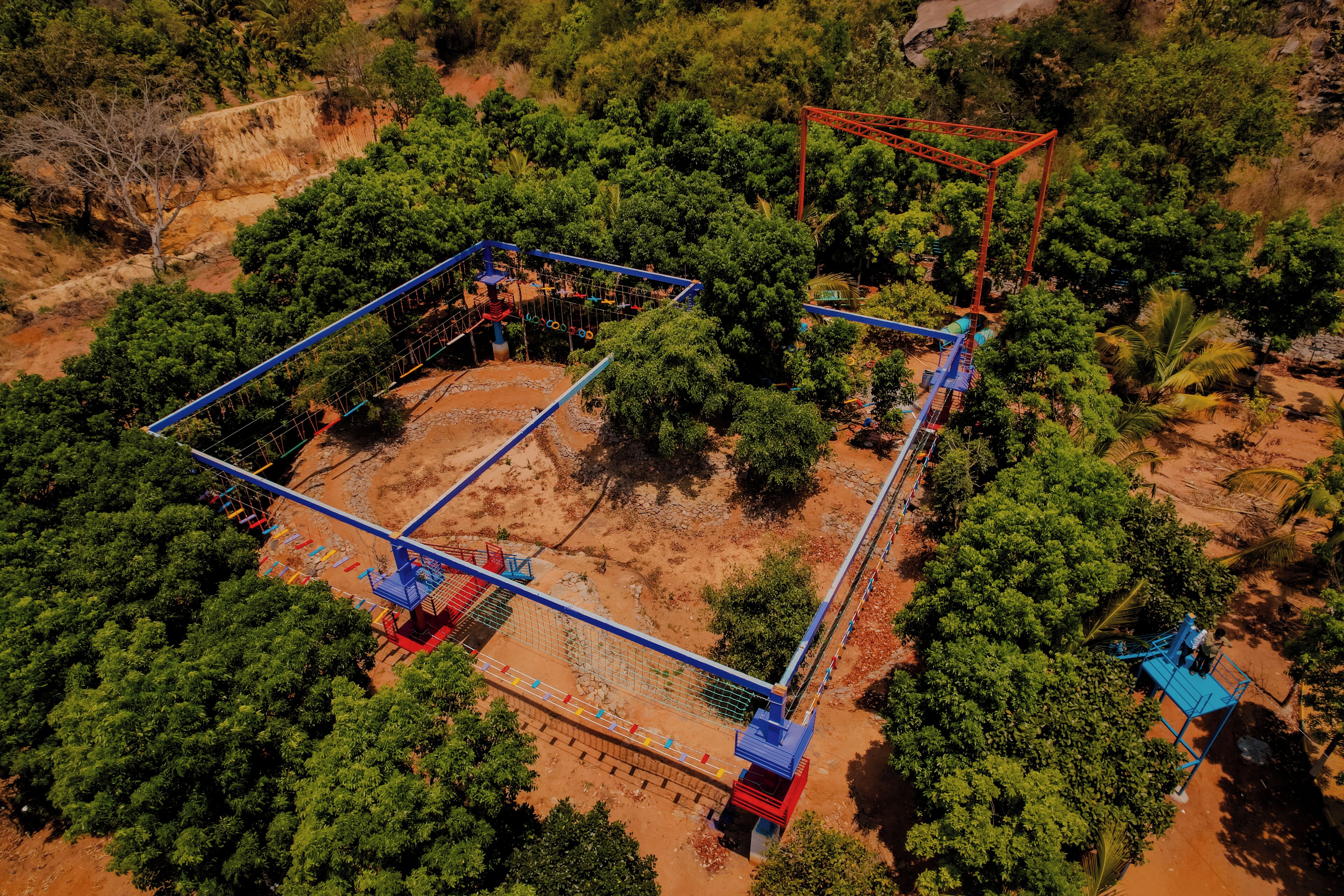 Vista aérea de un parque con una estructura de juego azul y roja, rodeada de frondosas plantas verdes y suelo de tierra.