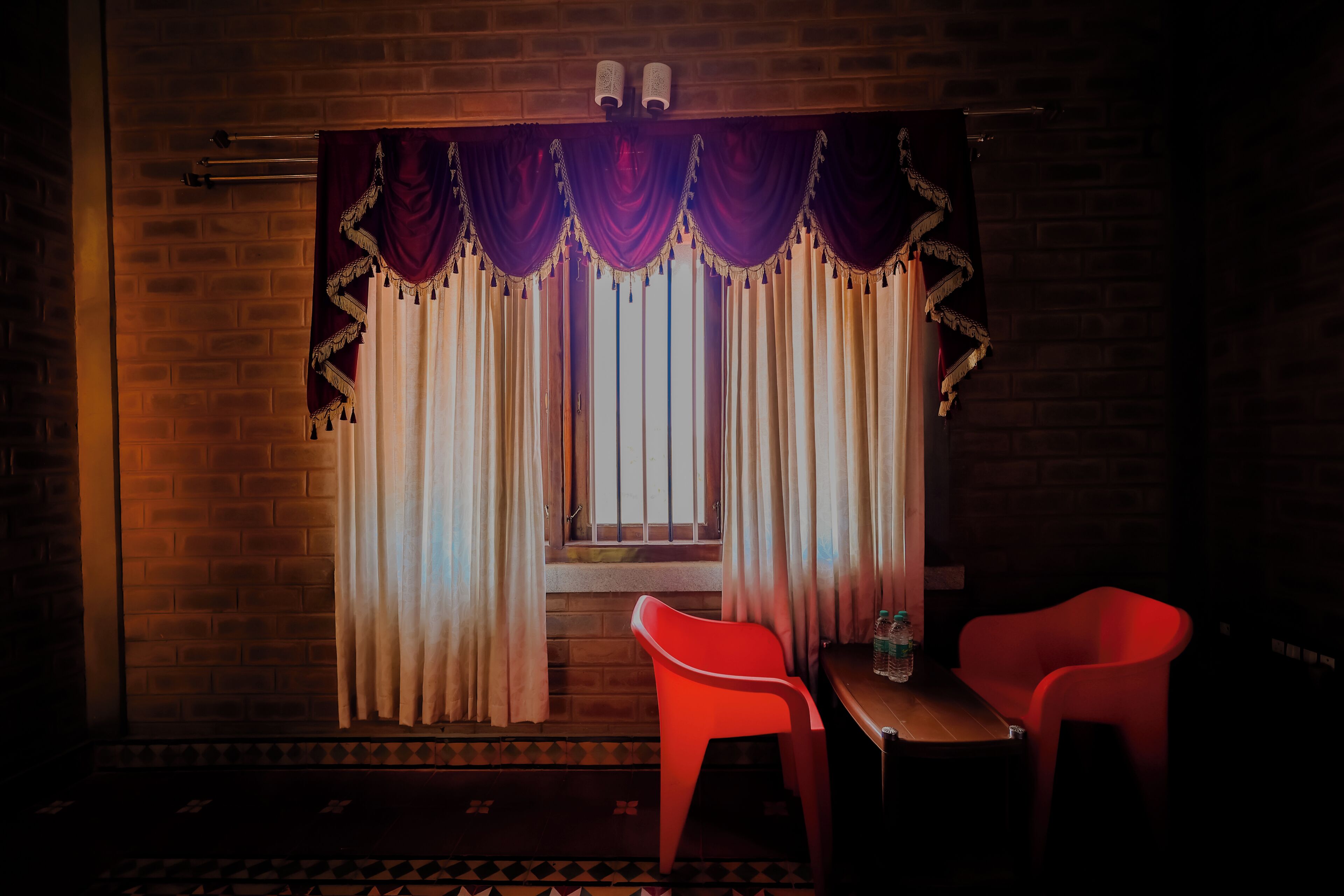 A room with a brick wall, a window with purple and white curtains, and two red chairs.