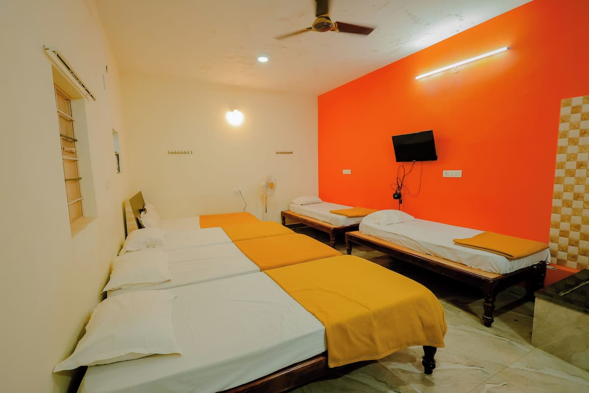 Habitación de hotel con tres camas individuales, un ventilador de techo, un televisor montado en una pared naranja y suelo de baldosas.