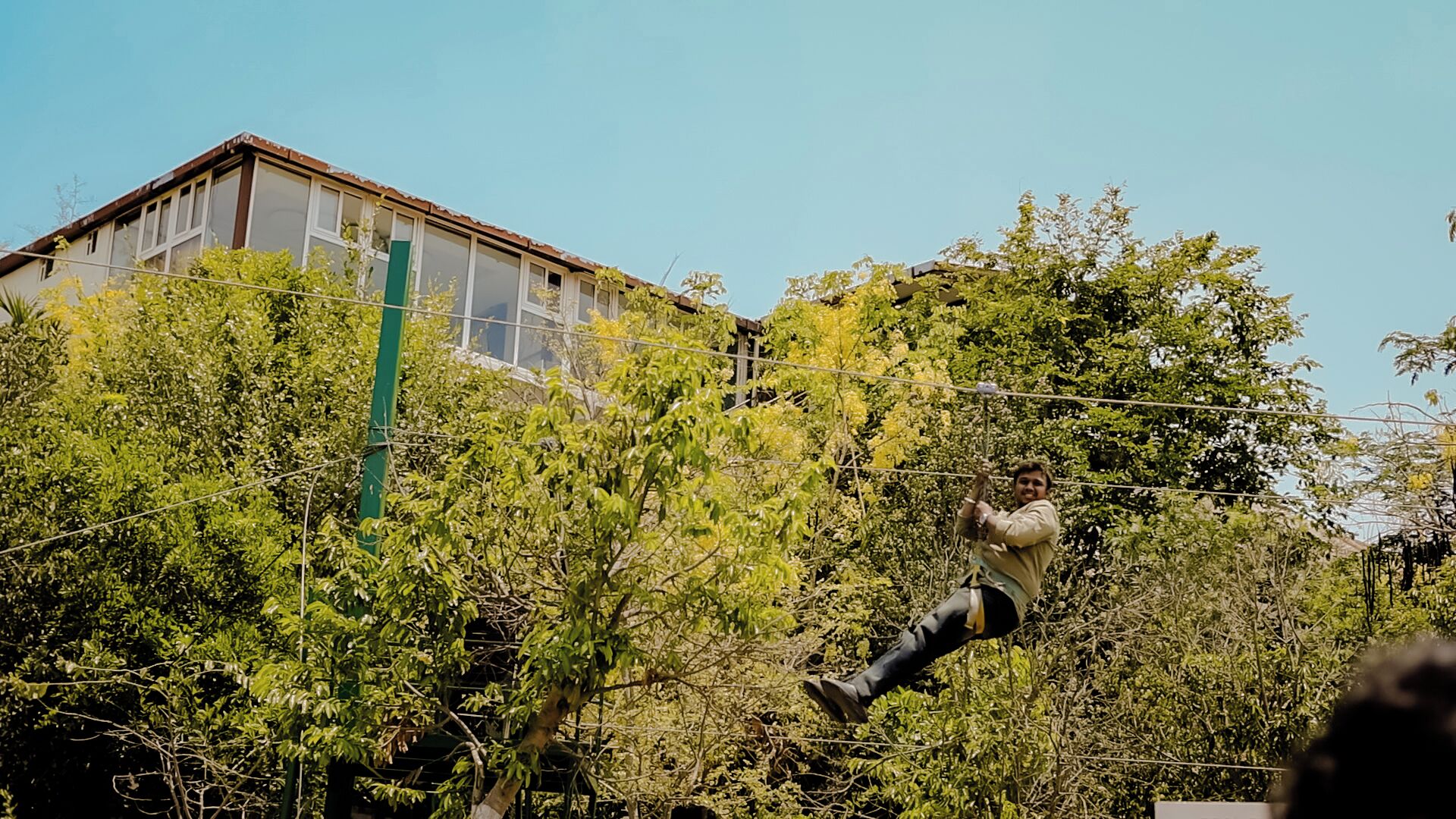 Uma pessoa executando um número de acrobacia em uma corda, cercada por vegetação densa e um edifício ao fundo.