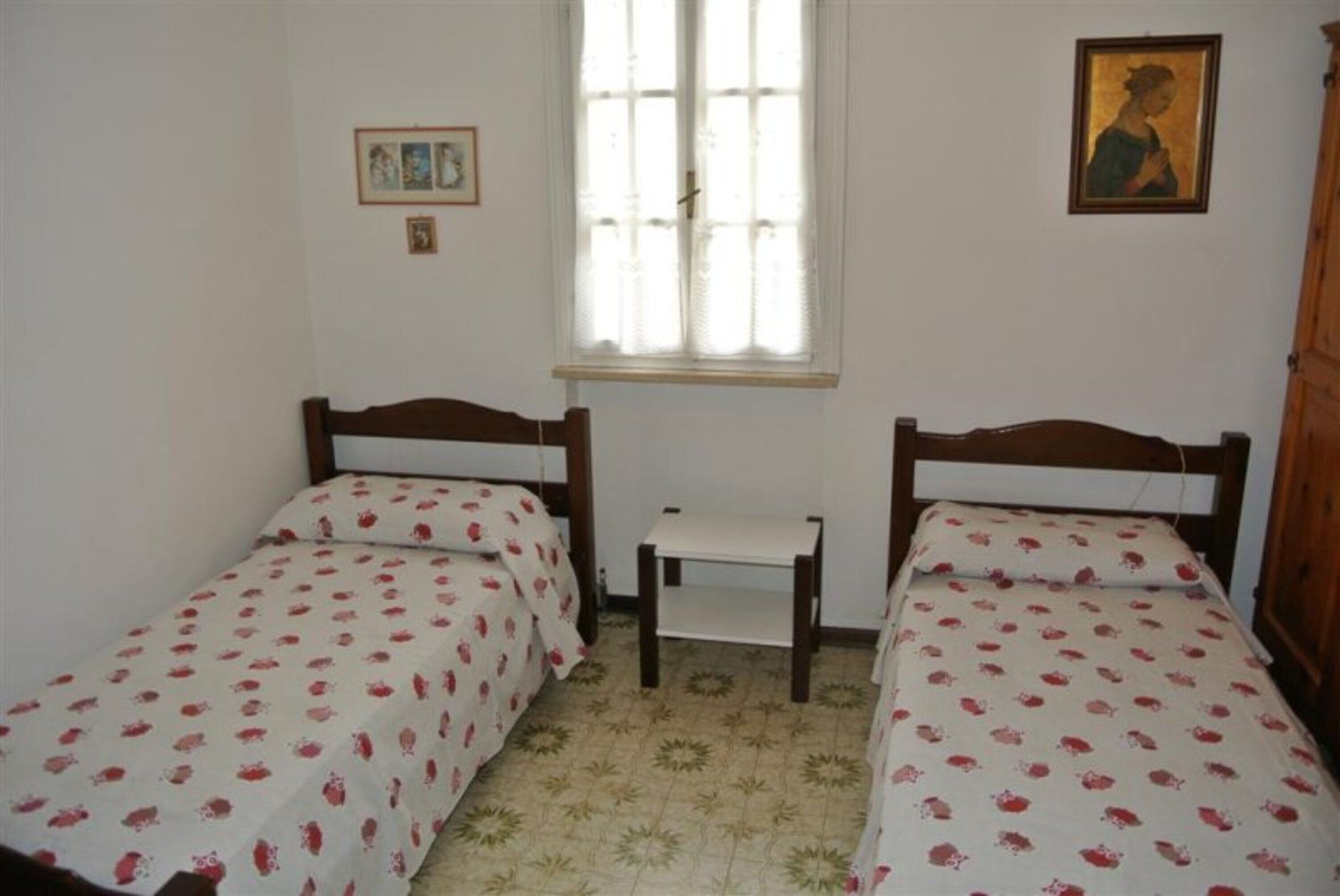 A room with two single beds, a small table, a window with curtains, and a framed picture on the wall.