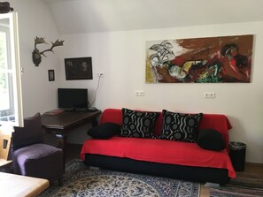 Ein Zimmer mit einem roten Sofa, gemusterten Kissen, einem Schreibtisch mit einem Computer, einem gerahmten Bild und einer Wanddekoration mit einem Hirschkopf.