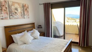 Ein Schlafzimmer mit einem großen Bett, einem Nachttisch, einer Lampe, Blick auf die Stadt durch eine Schiebetür und einem Gemälde an der Wand.