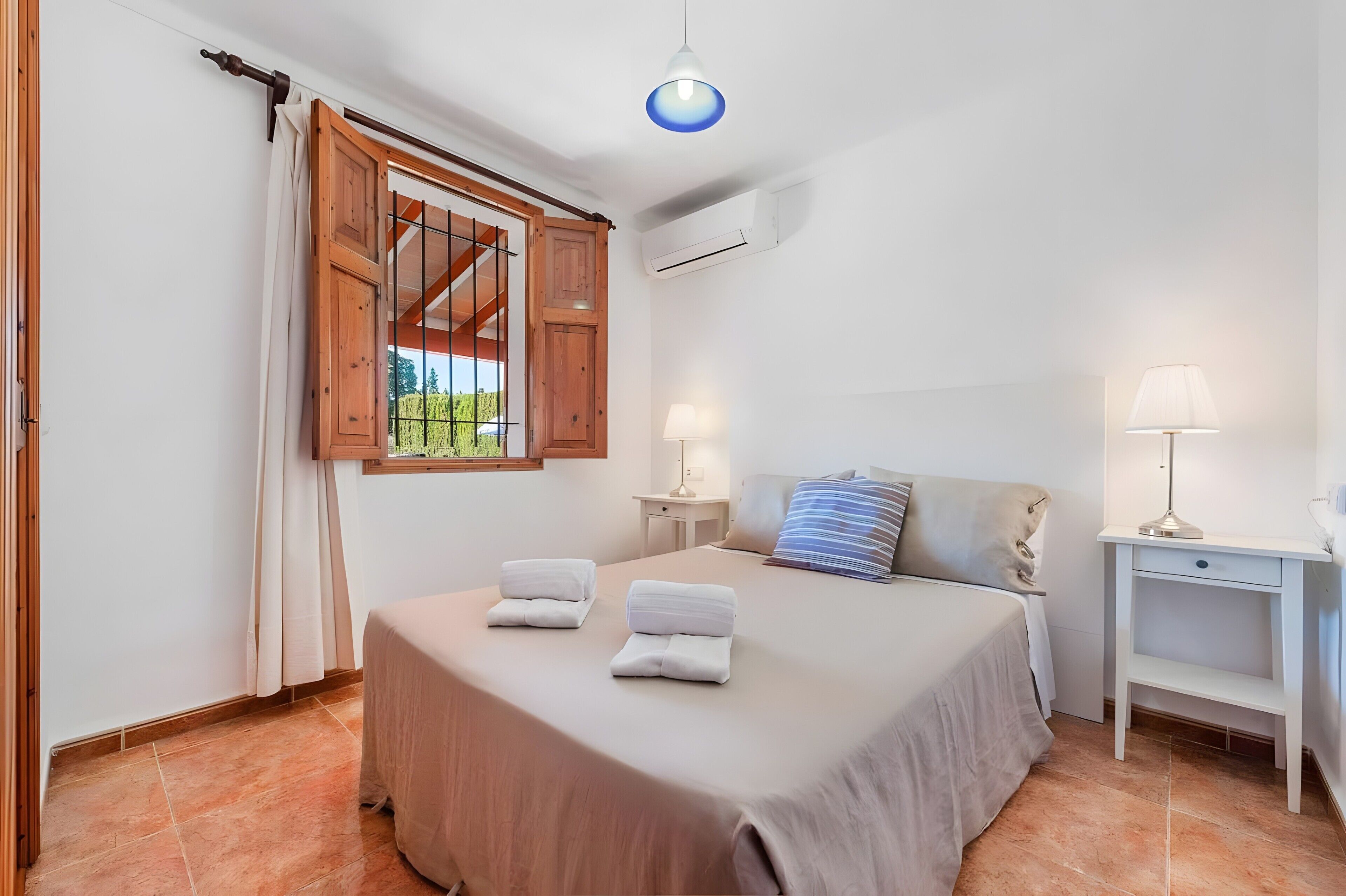 A bedroom with a bed, two bedside tables, a window with curtains, and an air conditioning unit.