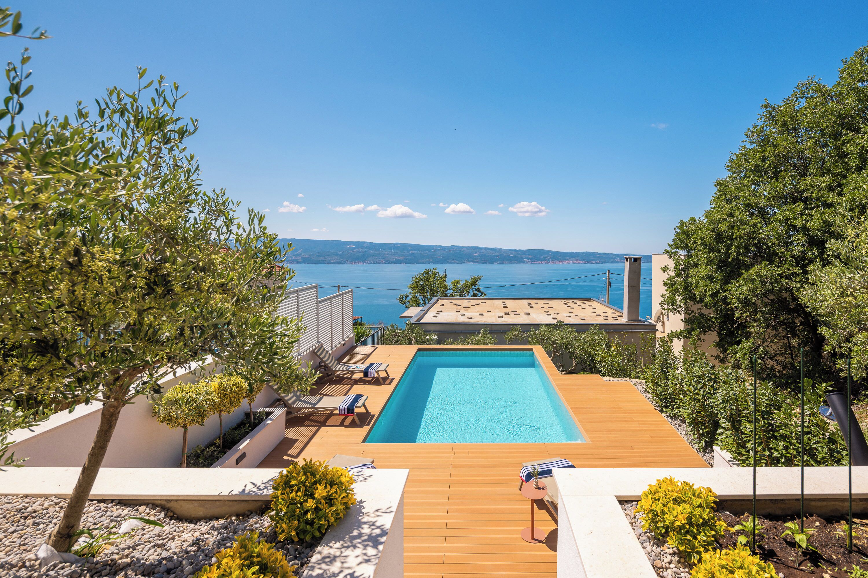 A pool with a wooden deck, lounge chairs, and a view of the sea.