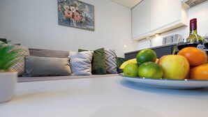 Ein modernes Wohnzimmer mit einer Couch, Kissen, einem Esstisch mit Obst und einer Flasche sowie einer Küchenzeile mit Schränken und einem Gemälde.