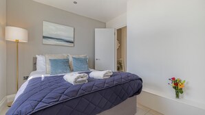 Ein Schlafzimmer mit Bett, Kissen, einer Nachttischlampe, einem Gemälde, einer Tür und einer Vase mit Blumen.