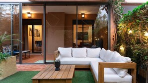 A modern outdoor living area with a wooden bench, a white sofa, and a glass door leading to an interior space.
