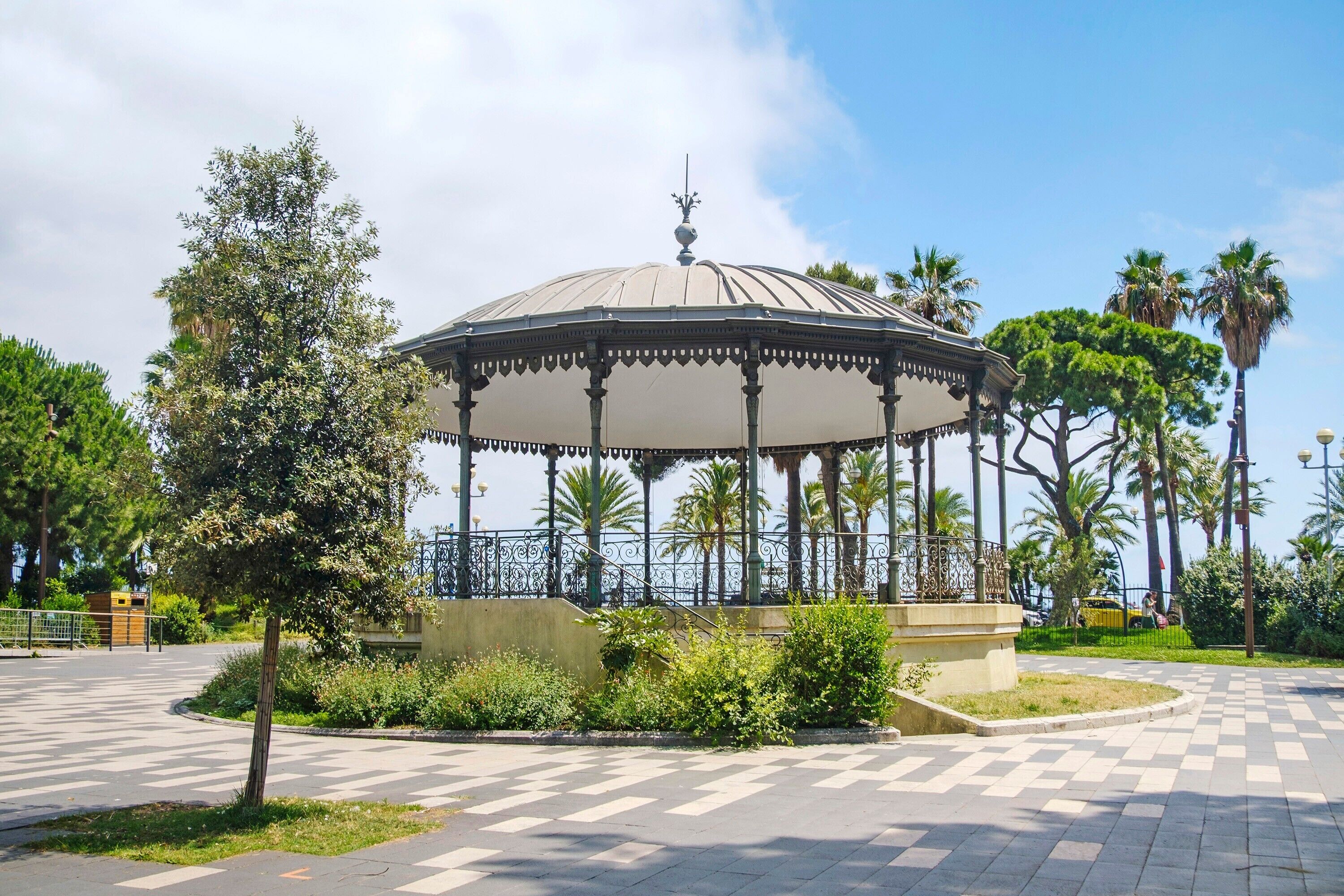 A gazebo with a dome roof, surrounded by a paved area with palm trees and a fence.