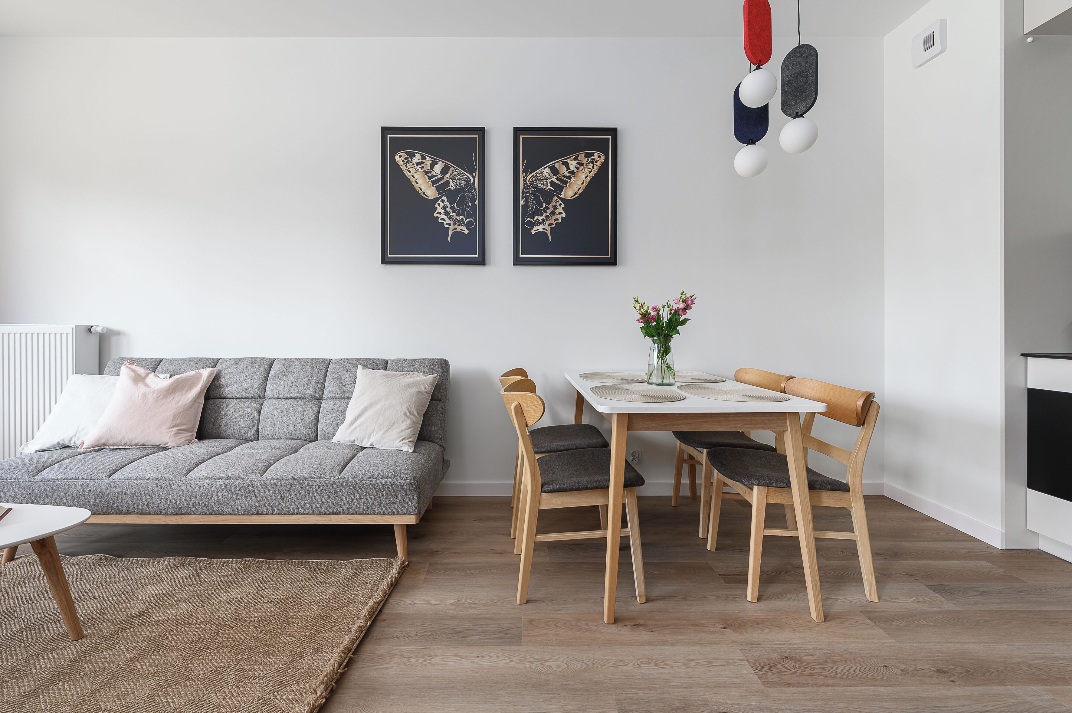 Ein modernes Wohnzimmer mit einem grauen Sofa, einem Esstisch mit Stühlen und zwei gerahmten Schmetterlingsbildern an der Wand.