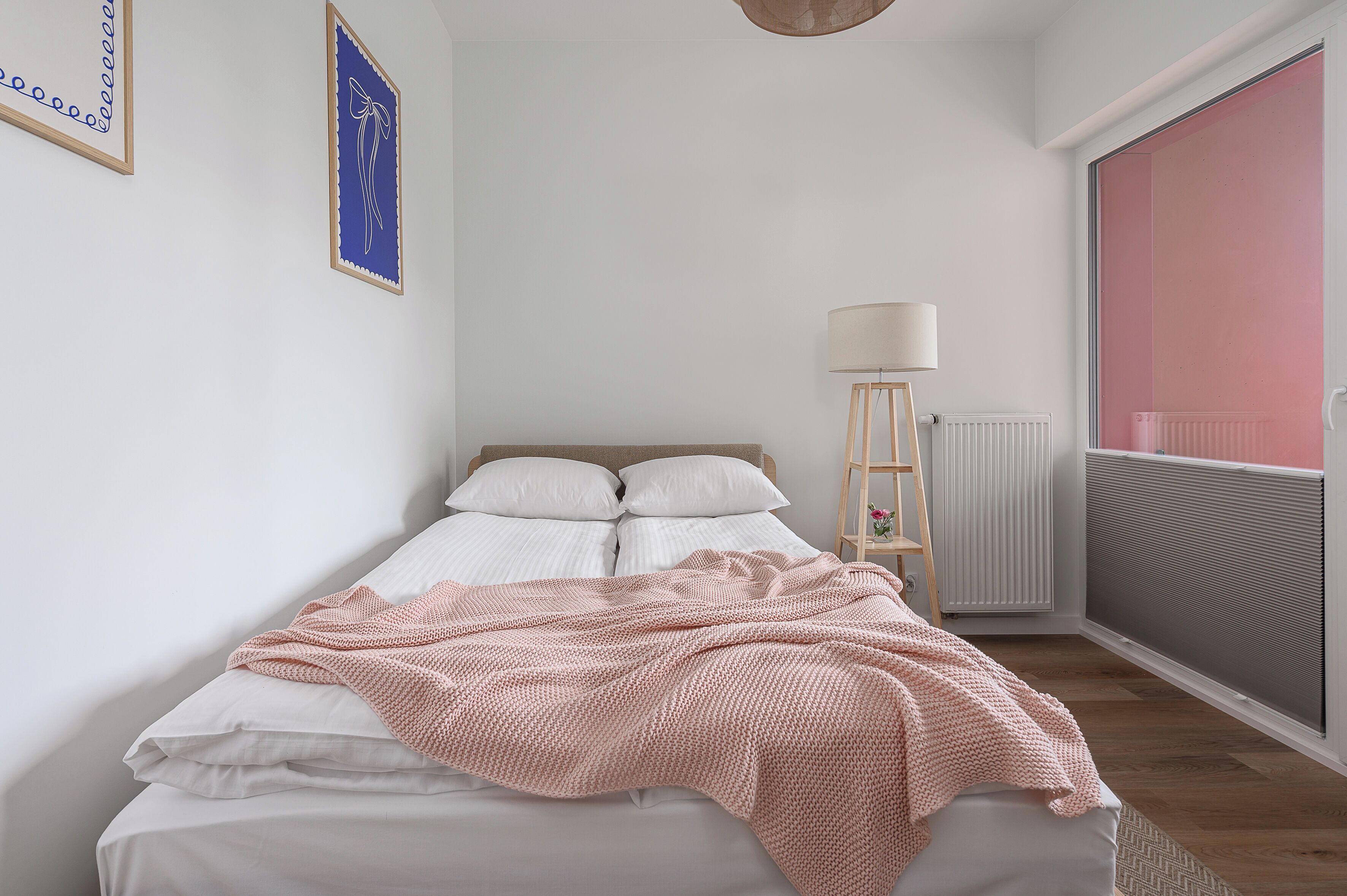 Ein Schlafzimmer mit Bett, Nachttischlampe, Heizkörper und Fenster mit Blick auf Rosa.