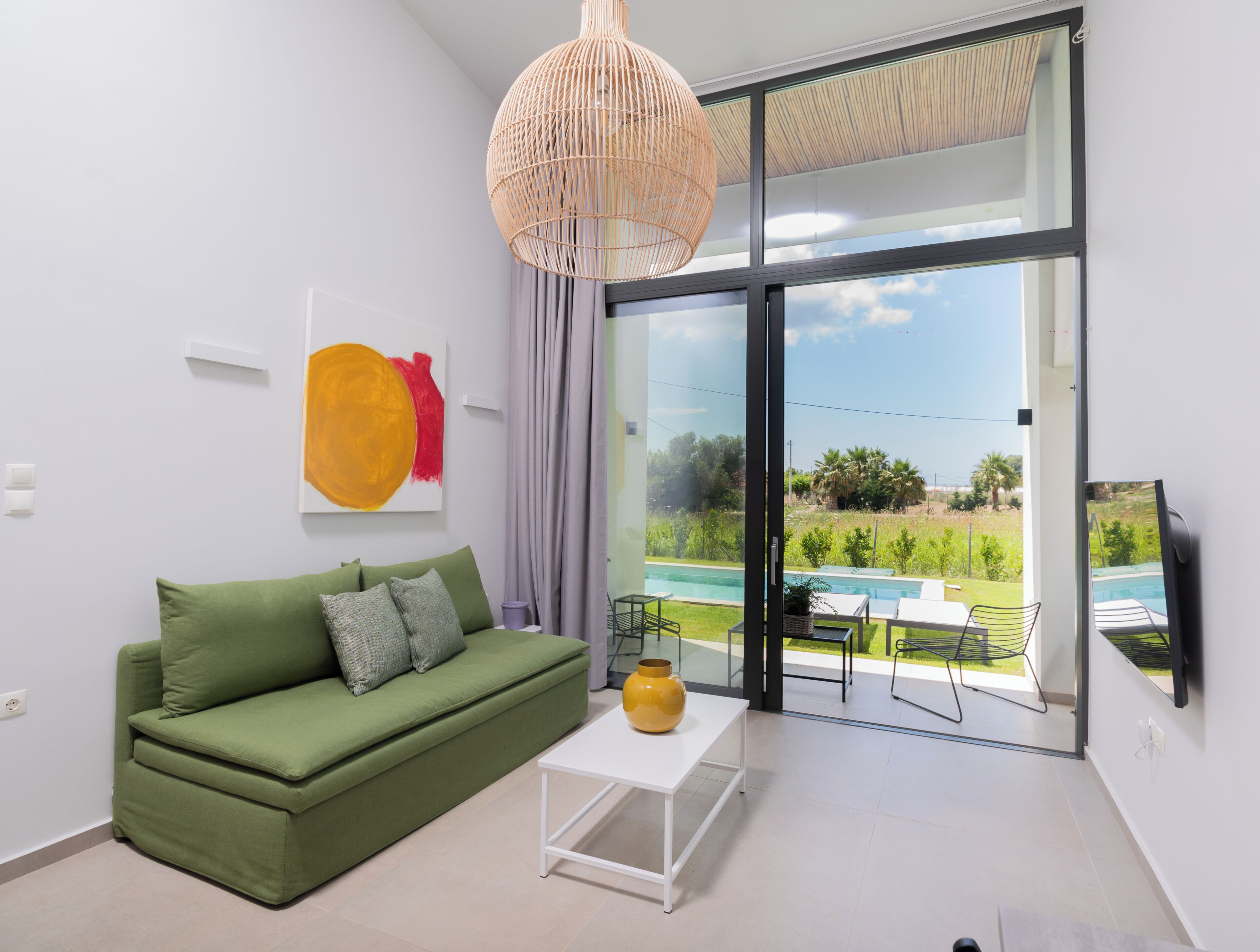 A modern living room with a green sofa, a white coffee table, a yellow vase, a large window with a view of a swimming pool and greenery.