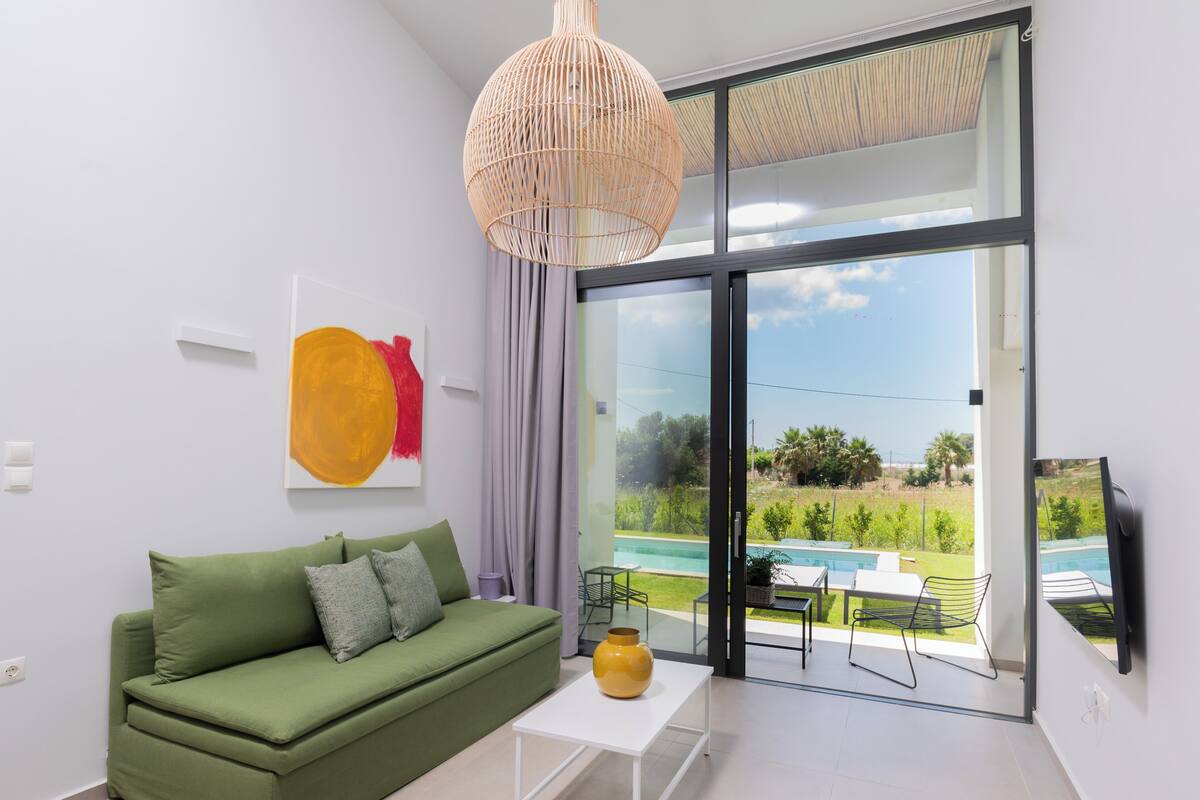 A modern living room with a green sofa, a white coffee table, a yellow vase, a large window with a view of a swimming pool and greenery.