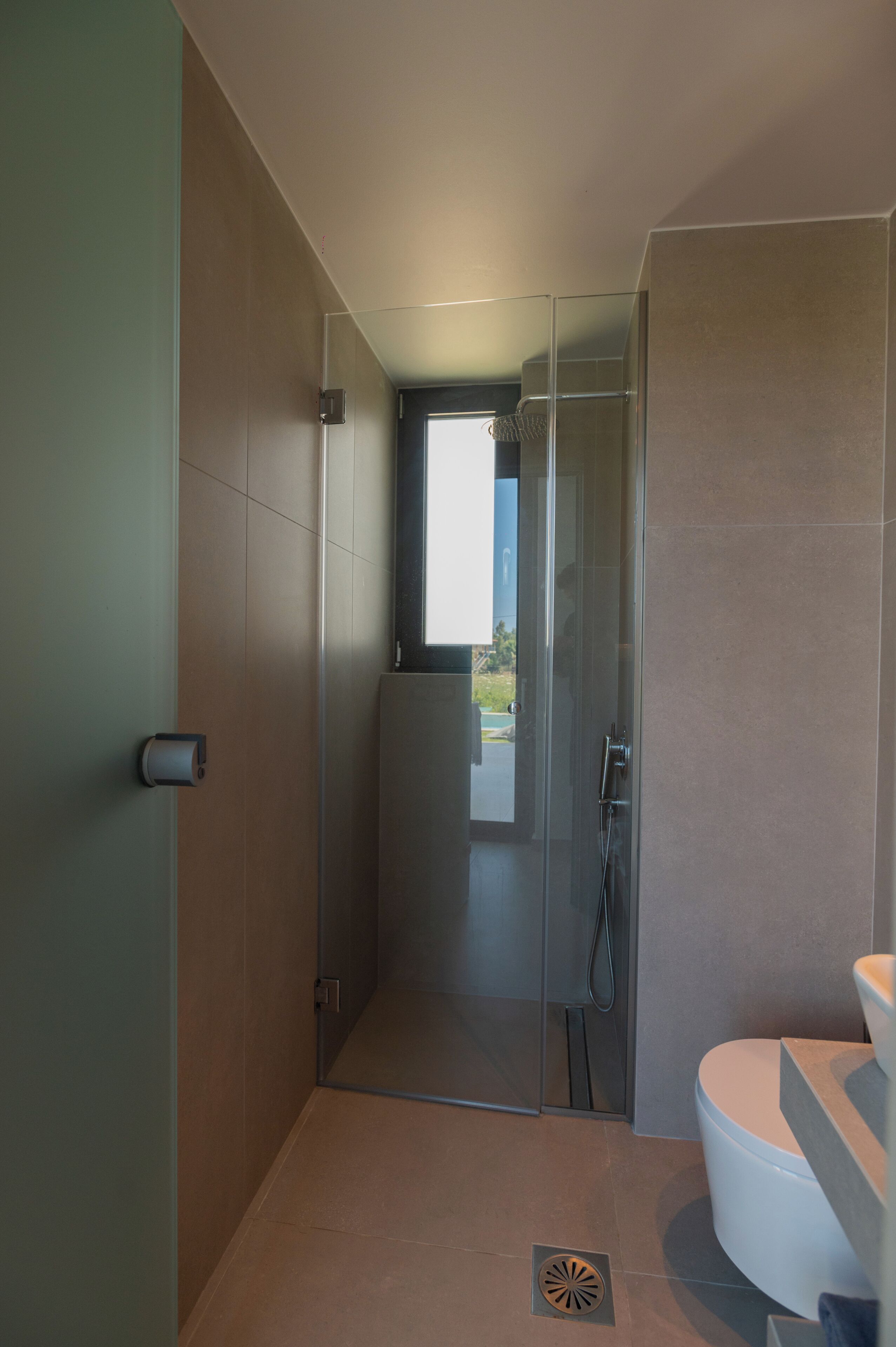 Casa de banho moderna com cabine de duche em vidro, sanita visível e porta que dá para uma varanda.