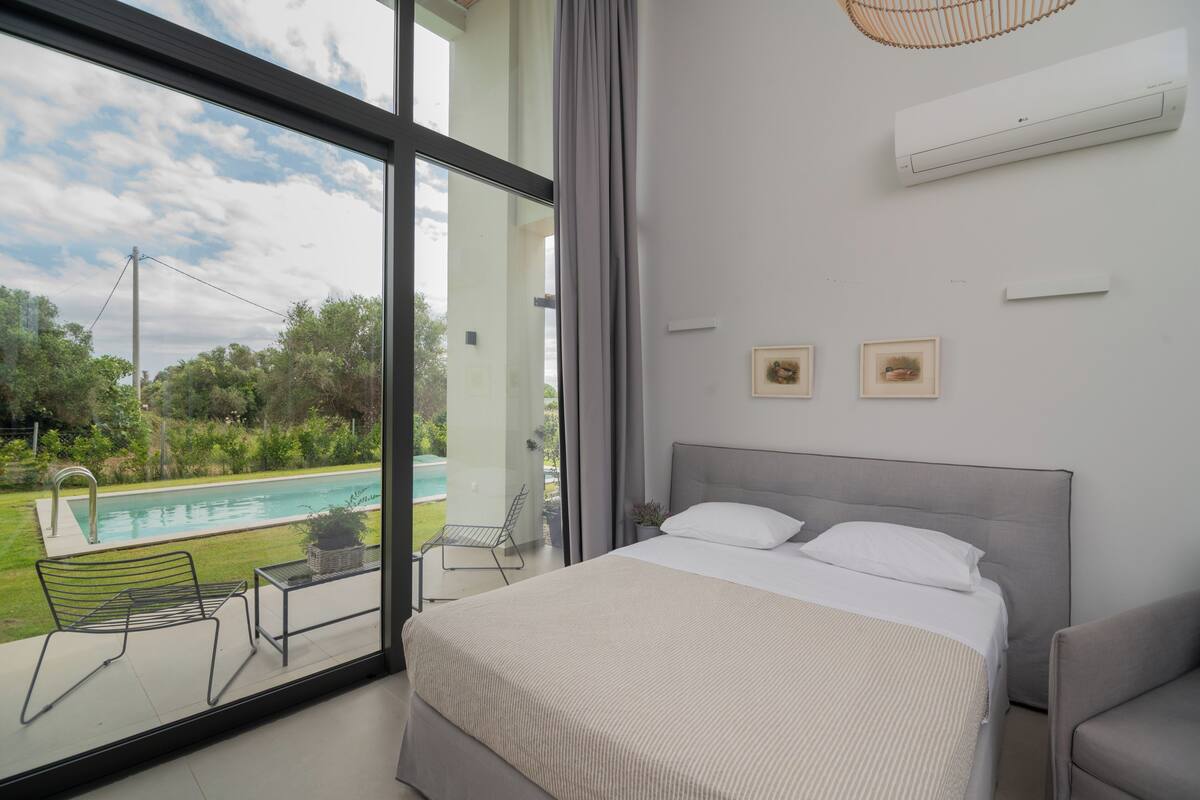 A bedroom with a large window, a bed, a chair, and a small table.